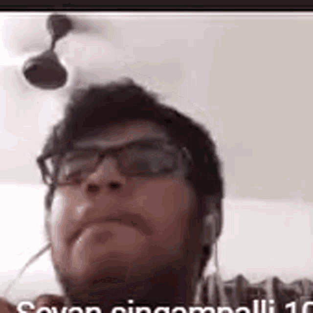 a man wearing glasses and headphones is talking on a video call with a ceiling fan in the background .