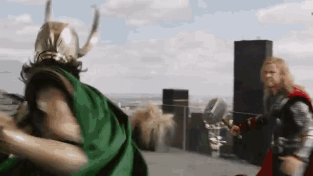 loki and thor are fighting in front of a city . thor is holding a hammer .