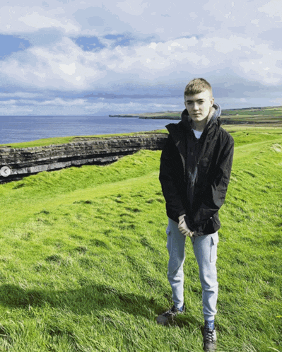 a boy in a black jacket stands in a grassy field