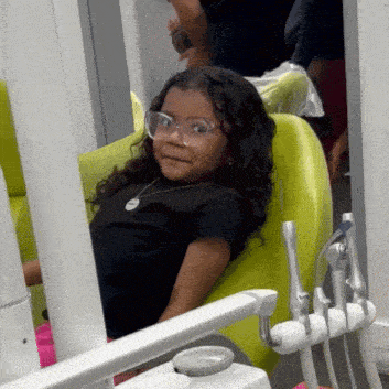a little girl wearing glasses and a black shirt is sitting in a dental chair