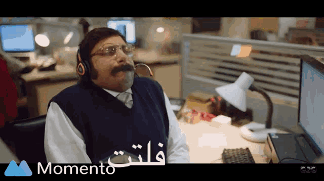 a man with a mustache wearing headphones sits at a desk with a momento logo on the bottom of the screen