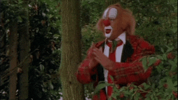 a clown in a red suit and tie is standing next to a tree in the woods .