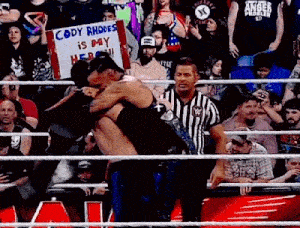a referee is standing in a wrestling ring holding a sign that says `` cody rhodes is my hero ''