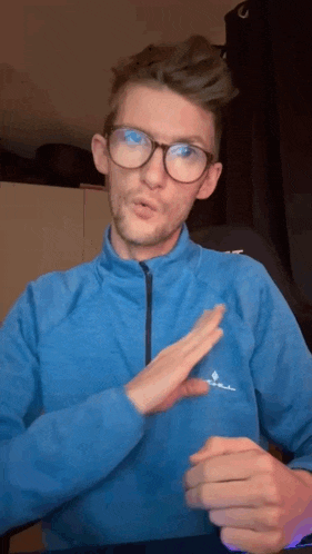 a man wearing glasses and a blue jacket is making a gesture with his hand .
