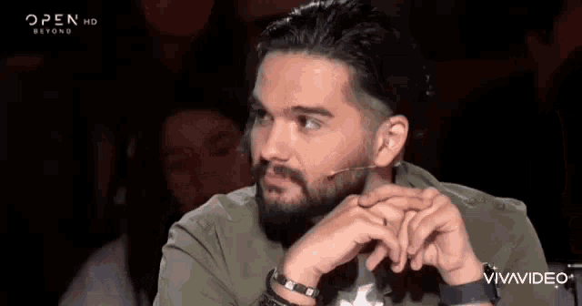 a man with a beard is sitting in front of a microphone with his hands on his chin .