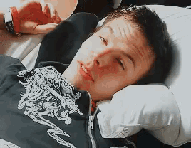 a young man is laying on a bed with his head on a pillow and wearing a black shirt with a dragon on it