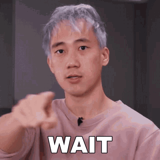 a man with gray hair is pointing at the camera with the word wait written on his chest .