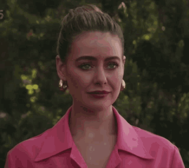 a woman wearing a pink shirt and gold hoop earrings stands in front of trees