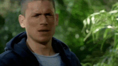 a man in a blue hoodie is standing in front of a tree and making a funny face .