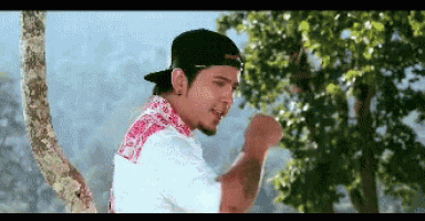 a young man wearing a baseball cap and a white shirt is standing in front of a tree .