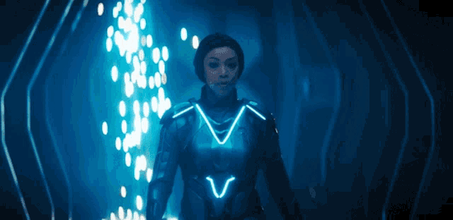 a woman in a futuristic suit is standing in a dark room with a blue light behind her