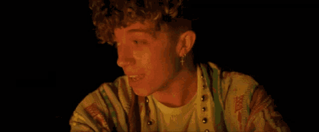 a young man with curly hair and earrings is sitting in front of a fire in the dark .
