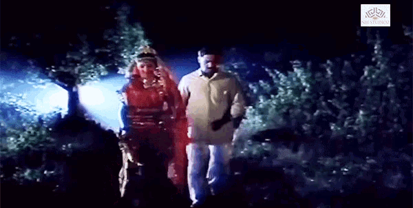 a man and a woman are walking through a dark forest with a logo for nv studios in the corner