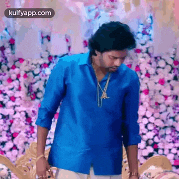 a man in a blue shirt and gold necklace is standing in front of a wall of flowers .
