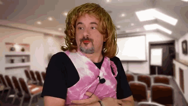 a man wearing a pink tie dye vest and a wig is sitting in a conference room .