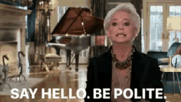 a woman is standing in a living room with a piano in the background and says " say hello be polite " .