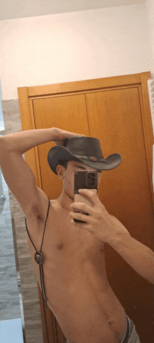 a shirtless man wearing a cowboy hat takes a selfie with his phone