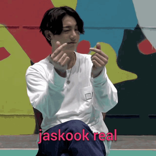 a man in a white shirt is making a heart shape with his hands and the word jaskook real is written above him