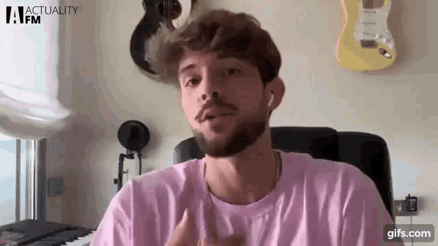 a man with a beard wearing a pink shirt is sitting in front of a microphone in a room .
