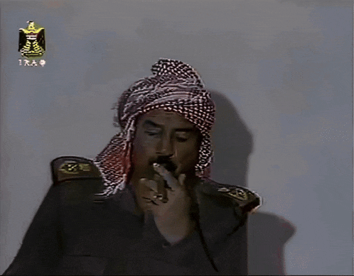 a man wearing a turban is smoking a cigarette in front of a wall that says iraq