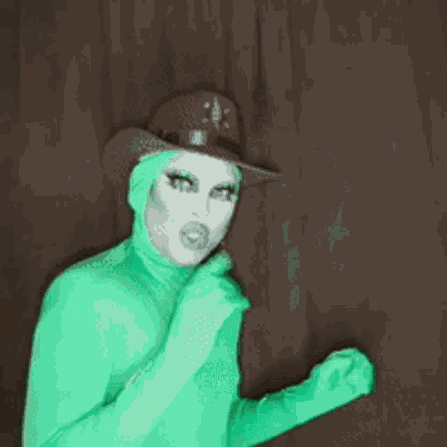 a drag queen wearing a cowboy hat and green gloves giving a thumbs up