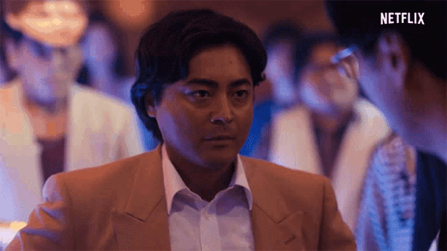 a man in a tan suit and white shirt is standing in a crowd of people .