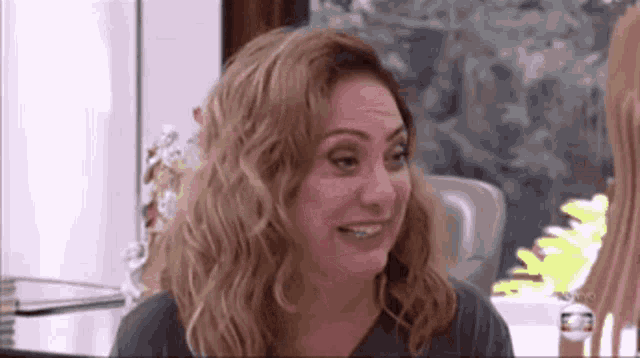 a woman is smiling and looking at the camera while sitting at a table in front of a window .
