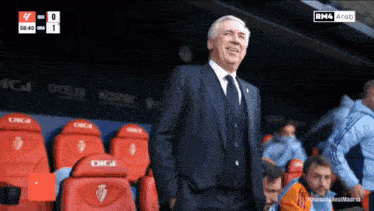 a man in a suit and tie stands in a stadium with red seats that say digi