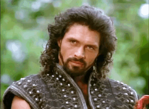a man with long hair and a beard is wearing a studded vest and looking at the camera .