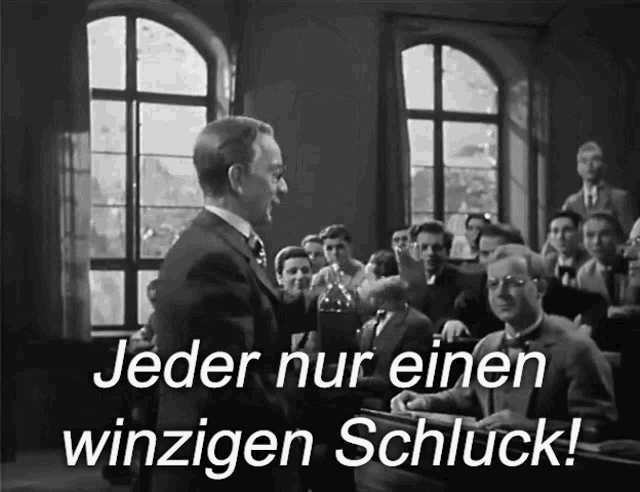 a man in a suit stands in front of a group of people and says jeder nur einen winzigen schluck