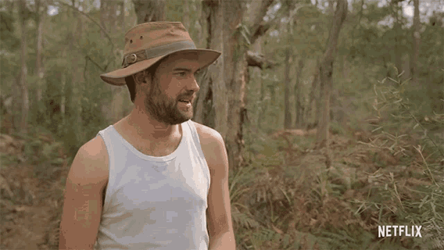 a man wearing a hat and a white tank top is standing in the woods with a netflix logo in the background