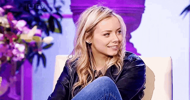a woman wearing a leather jacket and jeans is sitting in a chair and smiling .