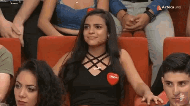 a woman wearing a black top with a red heart on it is sitting in a crowd of people watching a show on azteca uno