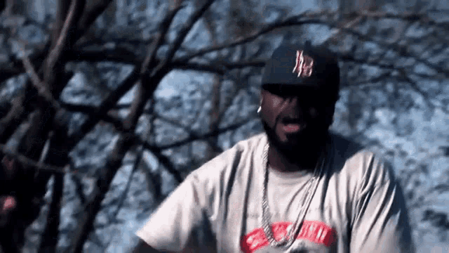 a man wearing a hat and a chain is standing in front of a tree .