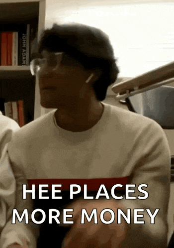 a man wearing glasses and ear buds is sitting in front of a bookshelf and saying hee places more money .