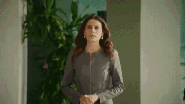 a woman in a grey jacket and earrings is standing in a hallway with her hands folded .