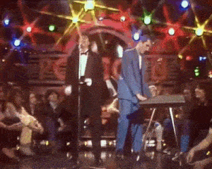 a man in a blue suit is playing a keyboard while another man sings into a microphone on a stage .