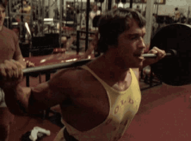 arnold schwarzenegger is squatting with a barbell in his mouth in a gym .