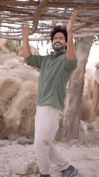 a man in a green shirt and white pants is standing under a wooden structure with his arms in the air .
