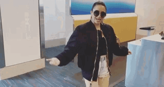 a woman wearing sunglasses and a bomber jacket is dancing in a room .
