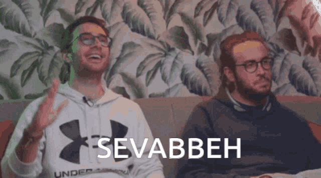 two men are sitting on a couch and one of them is wearing a white under armour hoodie