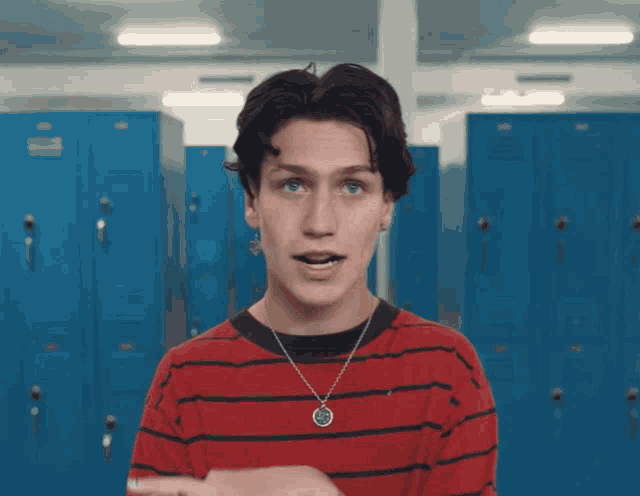 a young man wearing a red and black striped shirt and a necklace stands in front of blue lockers