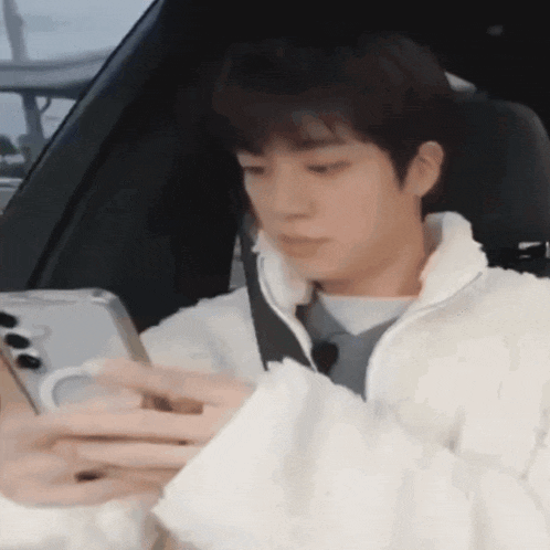 a man in a white jacket is sitting in a car looking at his phone .