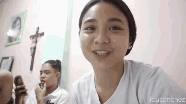 two girls are sitting in a room with a cross on the wall . one girl is smiling and the other is looking at her phone .