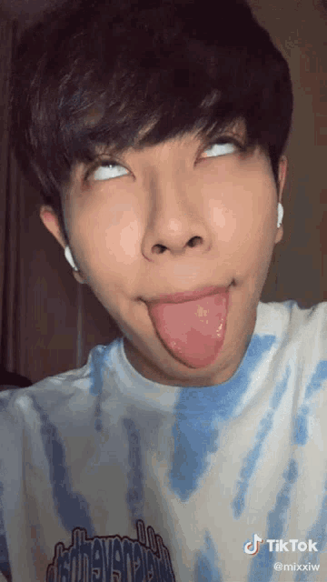 a young man wearing a blue tie dye shirt sticks his tongue out