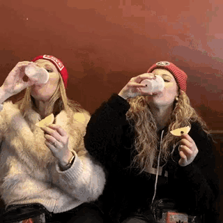 two women drinking from plastic cups while one wears a red hat that says coca cola
