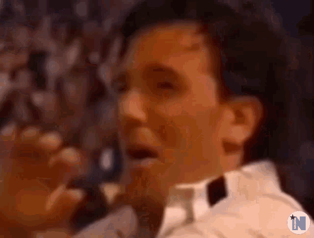 a man in a white shirt and black bow tie is singing into a microphone in front of a crowd .