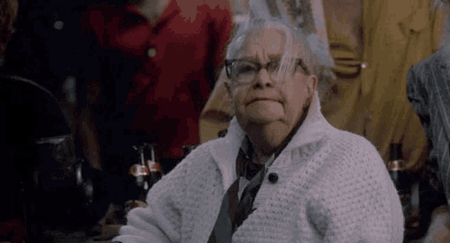an elderly woman wearing glasses and a white sweater is sitting at a table with bottles of soda .