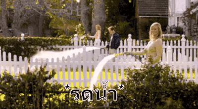 a woman is watering plants with a hose in front of a white picket fence in a garden .