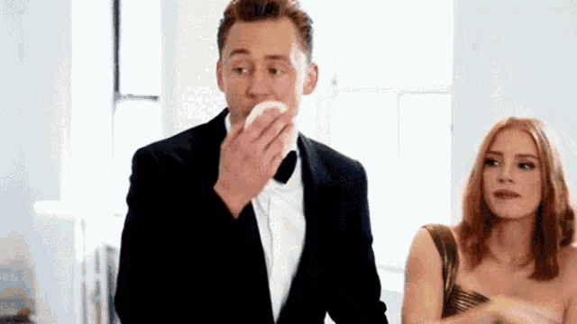 a man in a tuxedo is blowing his nose while a woman in a gold dress looks on .